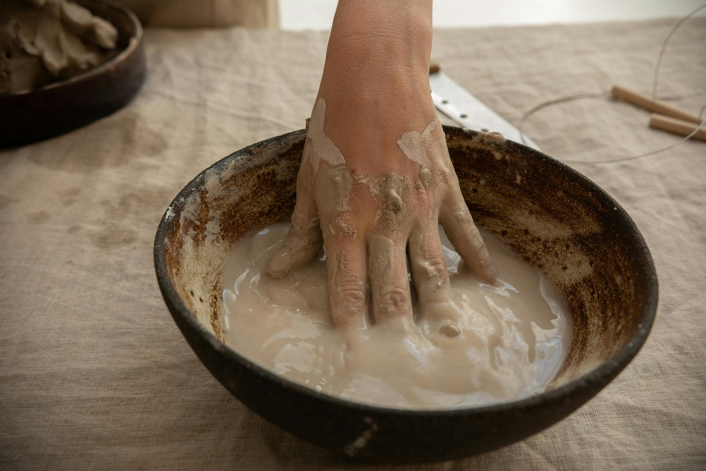 A hand mixing thick, grey clay in a dark ceramic bowl.