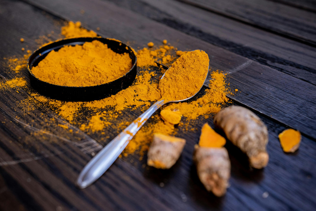 Bright yellow turmeric powder on a spoon and in a small bowl next to fresh turmeric roots.