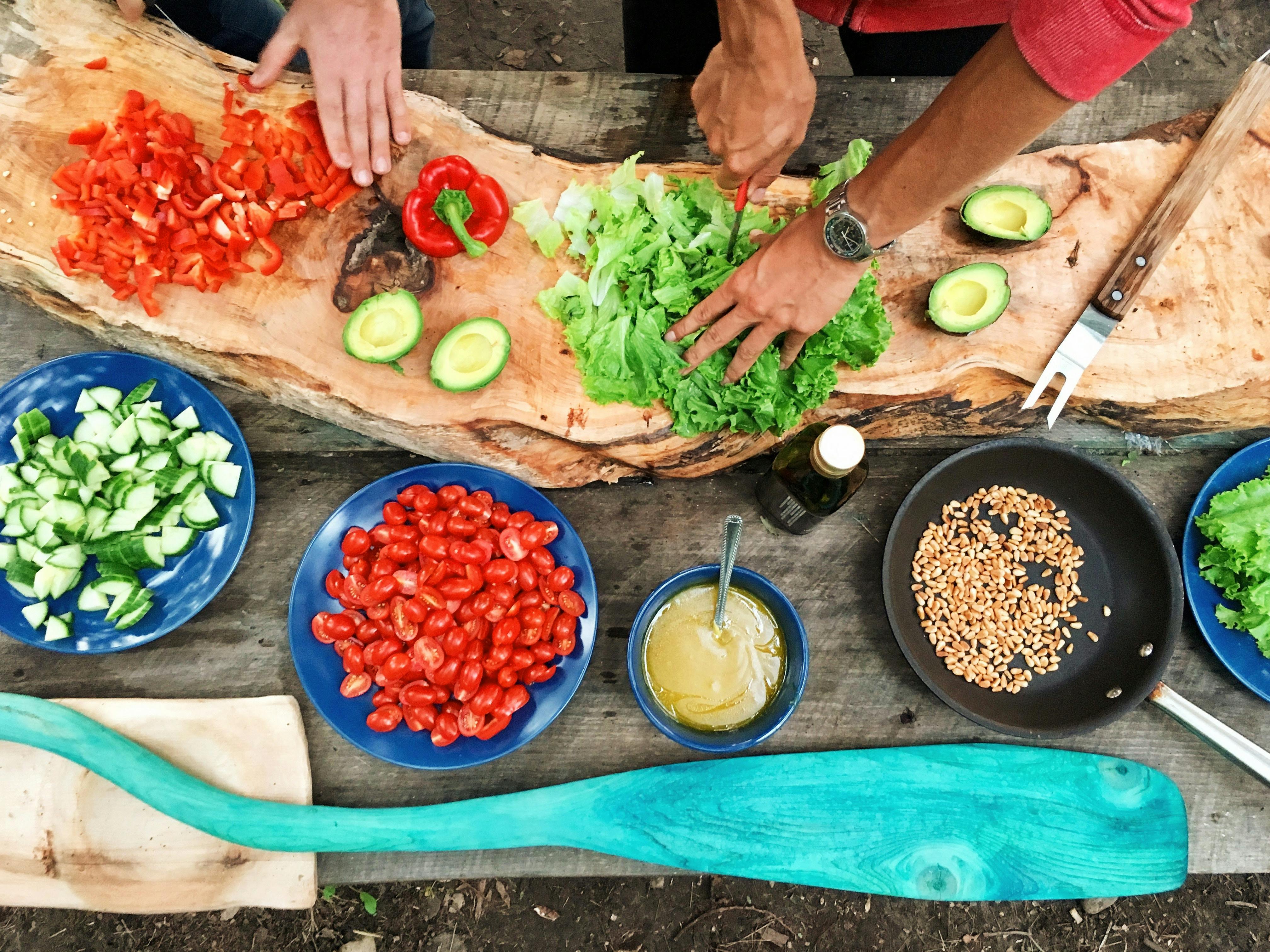 Top view of people chopping fresh vegetables like peppers, lettuce, and tomatoes on a large wooden board.