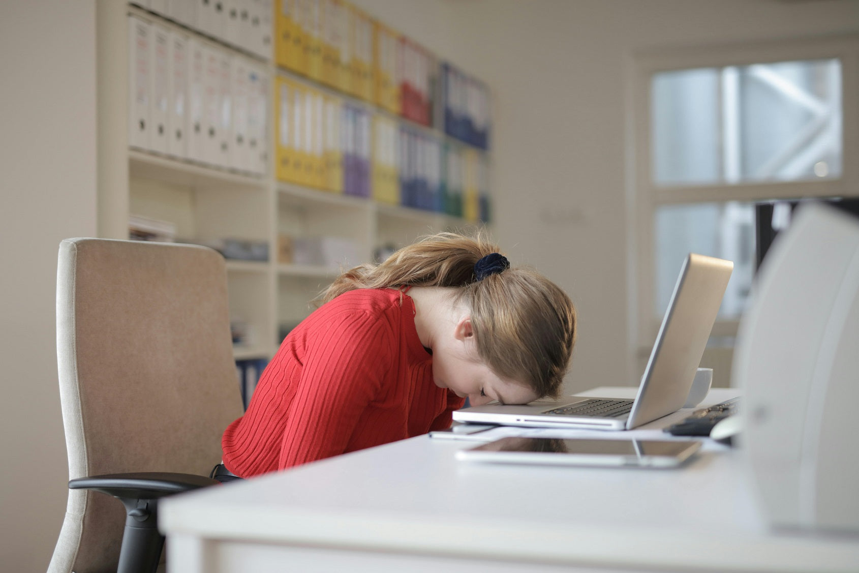A woman resting her head on her laptop keyboard in an office.