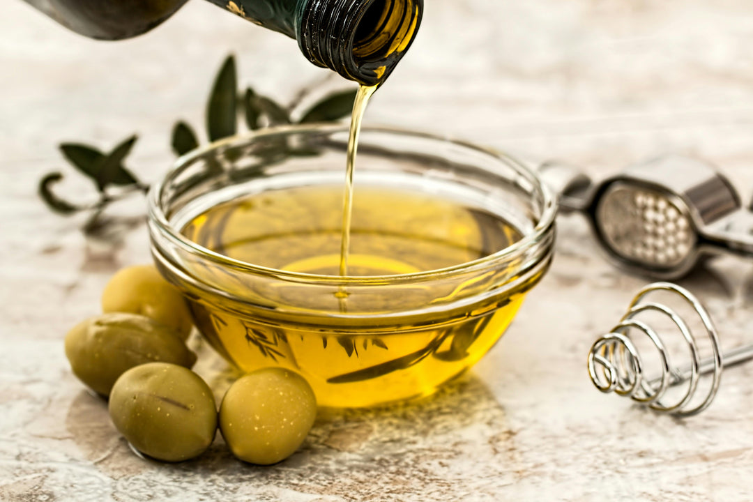 Olive oil pouring into a glass bowl with whole olives nearby.