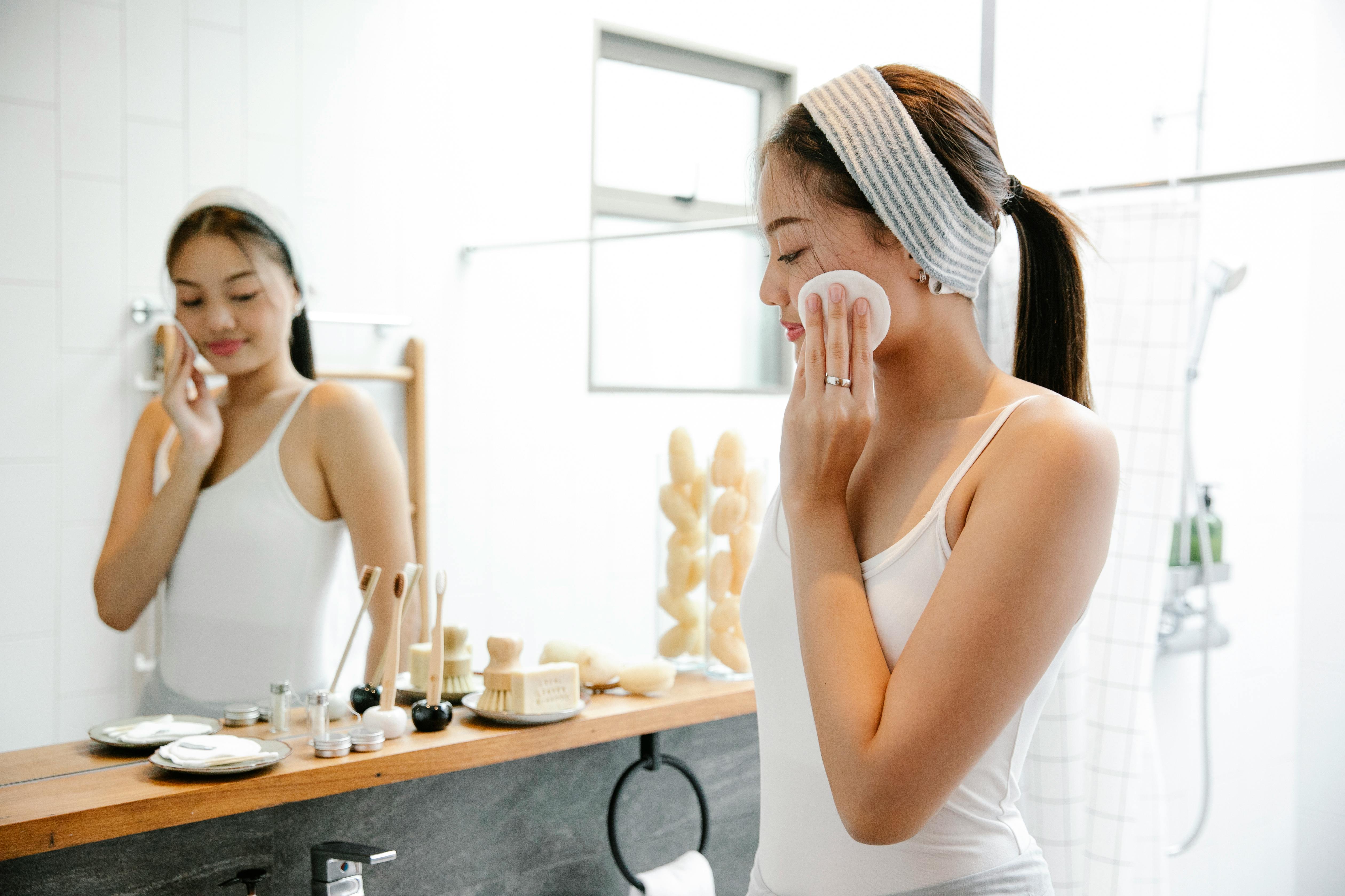 A woman cleaning her face with a cotton pad in front of a bathroom mirror.