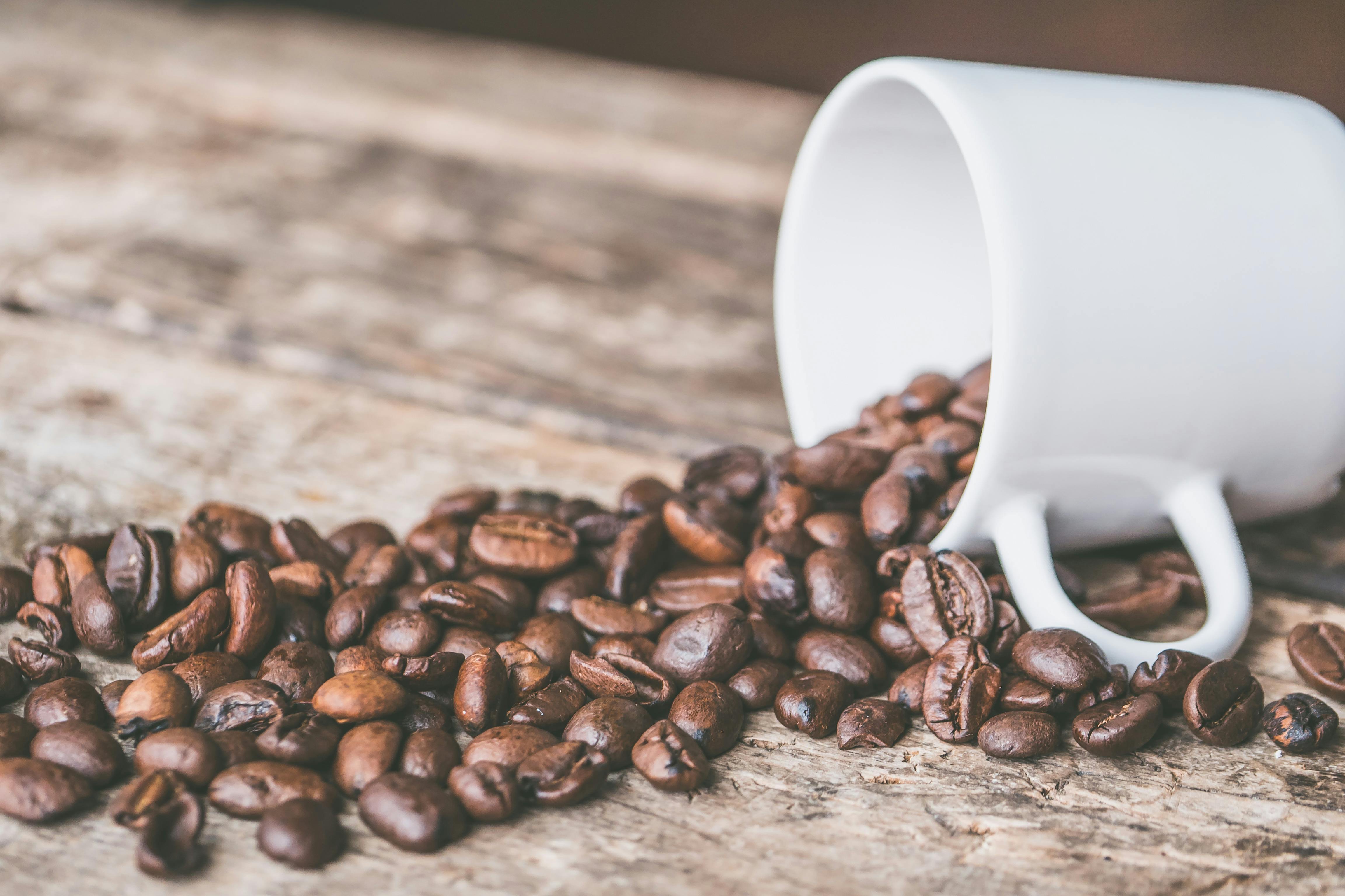 Roasted coffee beans spilling out of a white mug onto a wooden surface.